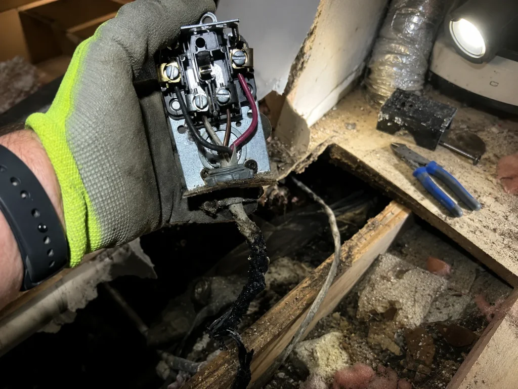 A Hayes Electrical technician holds a charred receptacle with melted Romex insulation pulled from fire-damaged subfloor framing, after-hours emergency response in Lake Charles.